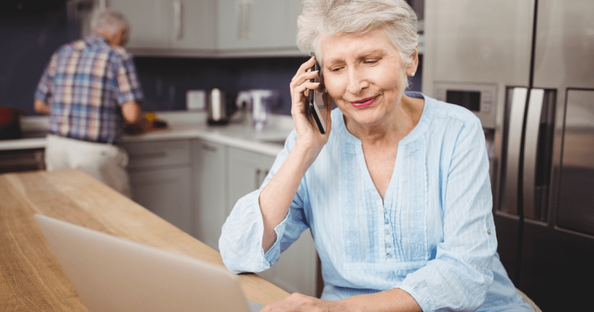An older woman talking on the phone while using a laptop for How seniors shop in the kitchen.